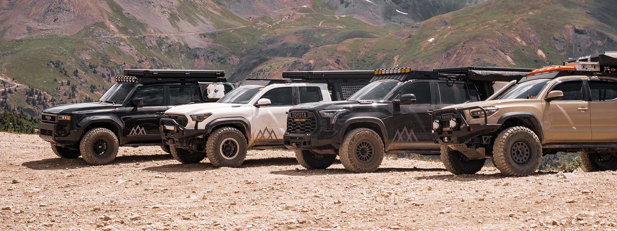 A 2024 toyota land cruiser, 2024 toyota tacoma, 2023 toyota tundra, and 2017 toyota tacoma with Backwoods Adventure Mods front bumpers parked next to each other on top of a mountain in colorado.