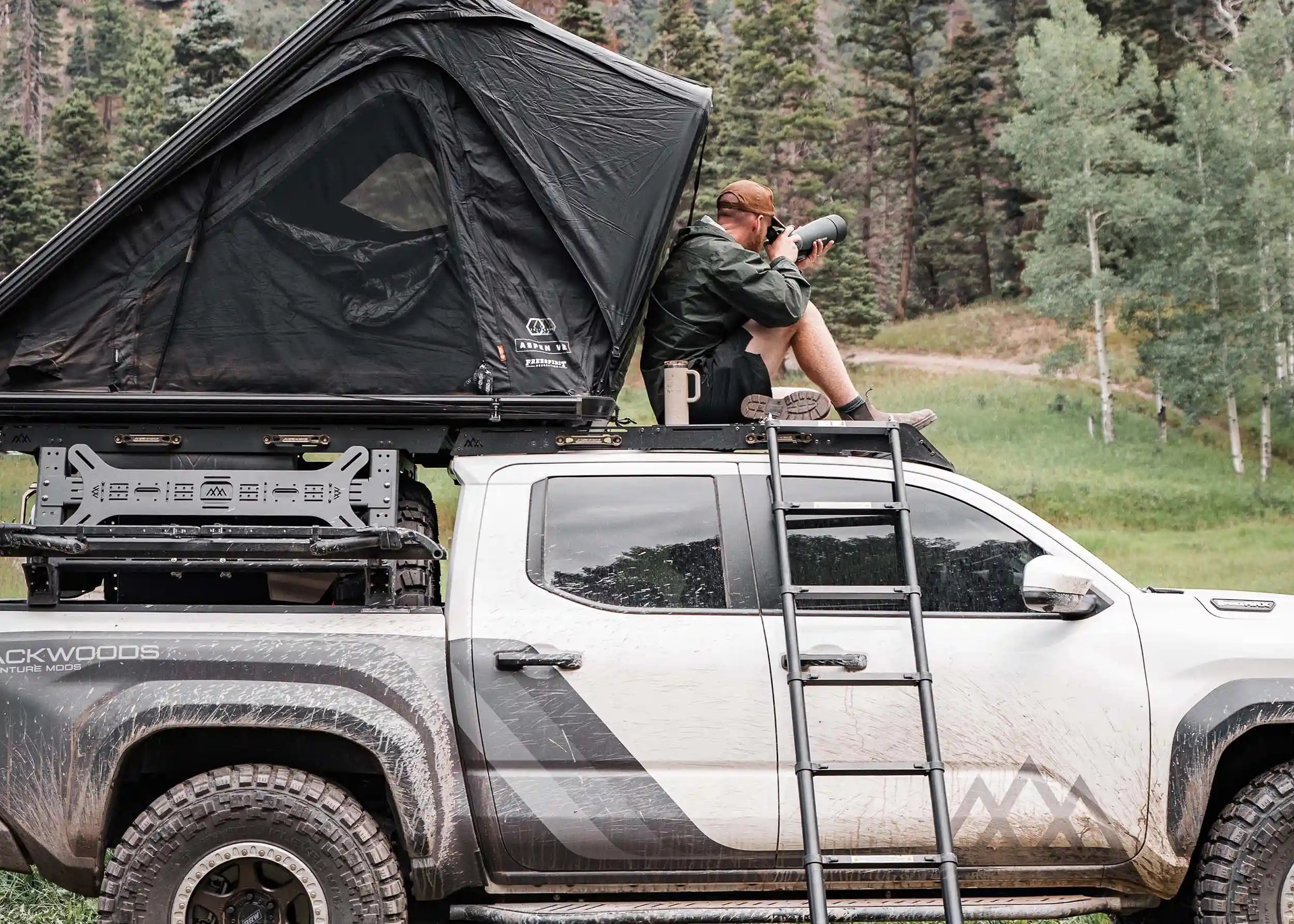 Man camping in a roof top tent mounted on top of a Backwoods Adventure Mods overland bed rack and roof rack installed on a 2024 4th gen Toyota Tacoma