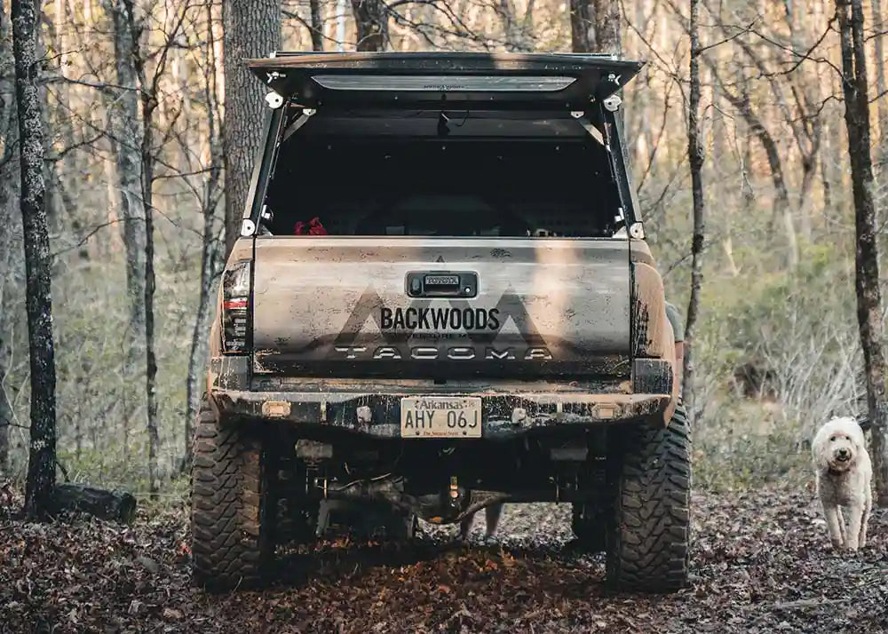 Rear-facing view of a 3rd Gen Toyota Tacoma overland camping off road with a Backwoods Adventure Mods Hi-Lite High Clearance Rear Bumper installed and covered in mud