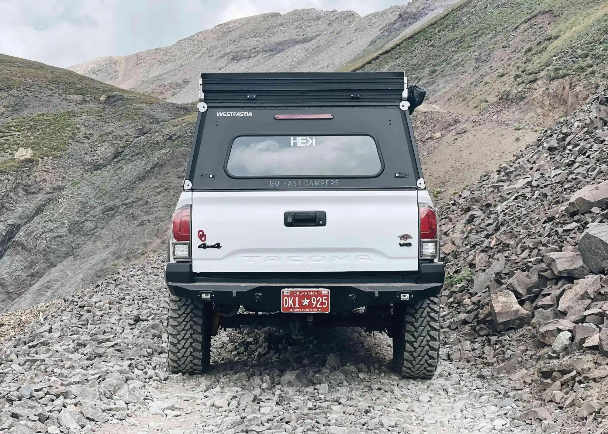 Rear-facing view of a White 3rd Gen Toyota Tacoma driving off road in the mountains with a Backwoods Adventure Mods Hi-Lite High Clearance Rear Bumper installed
