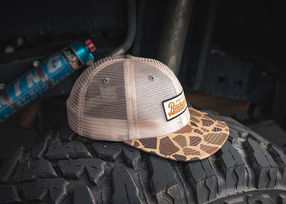 Backwoods Adventure Mods Camo Trucker hat baseball cap sitting on top of an off road vehicle tire