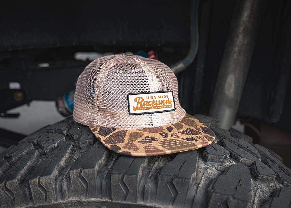 Backwoods Adventure Mods Camo Trucker hat baseball cap sitting on top of an off road tire