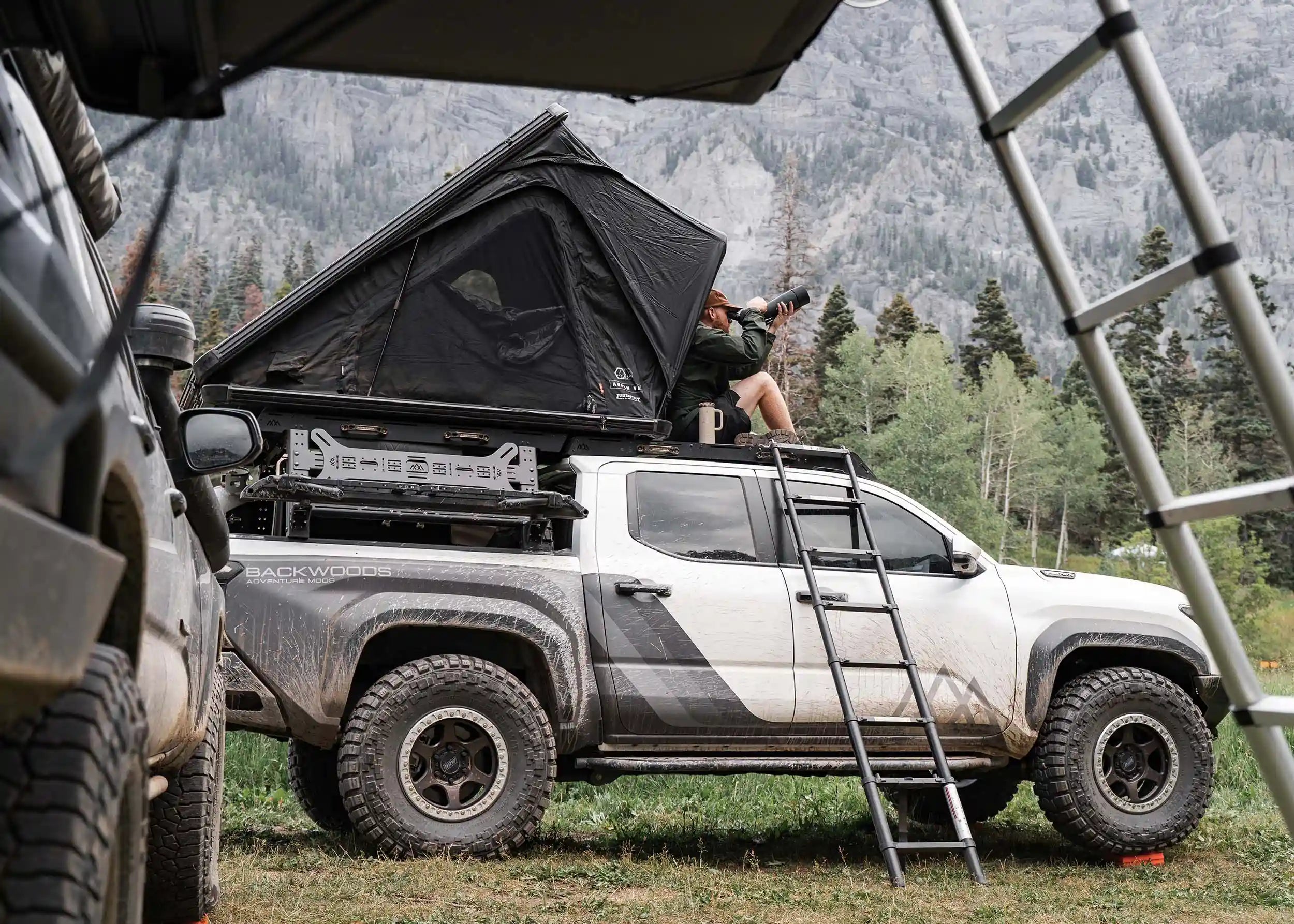 Man camping with the Backwoods Adventure Mods Toyota Tacoma Overland Bed Rack installed with roof top tent on top and off road accessories mounted to the sides