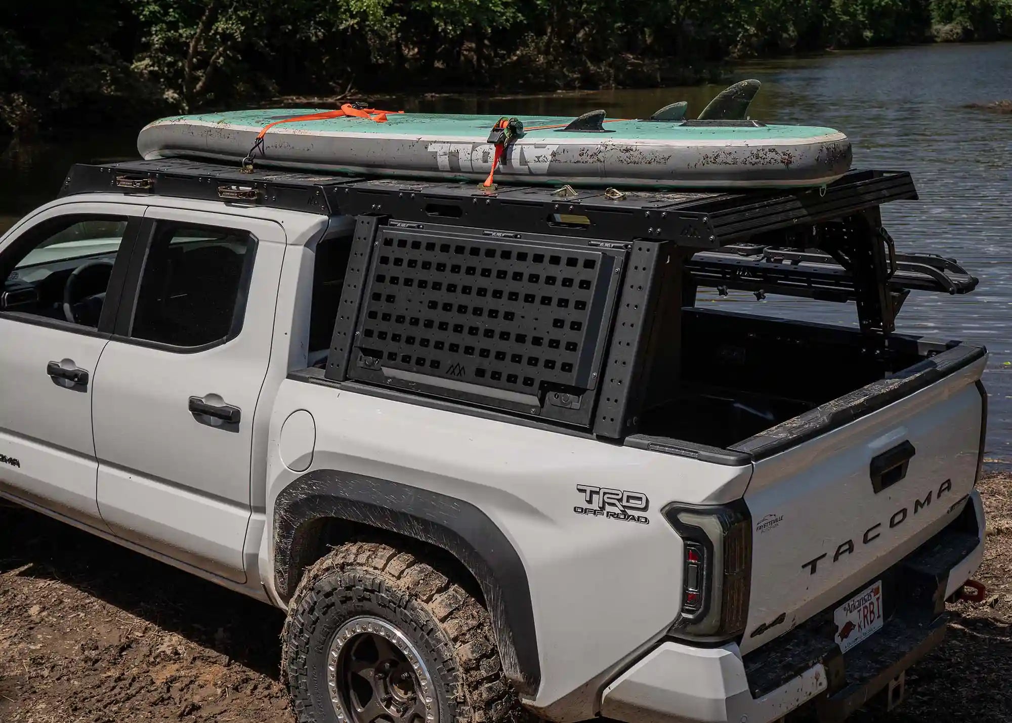 White Toyota Tacoma with a Backwoods Adventure Mods Tacoma Bed Rack and Tacoma Roof Rack combo hauling a paddleboard off road near a body of water