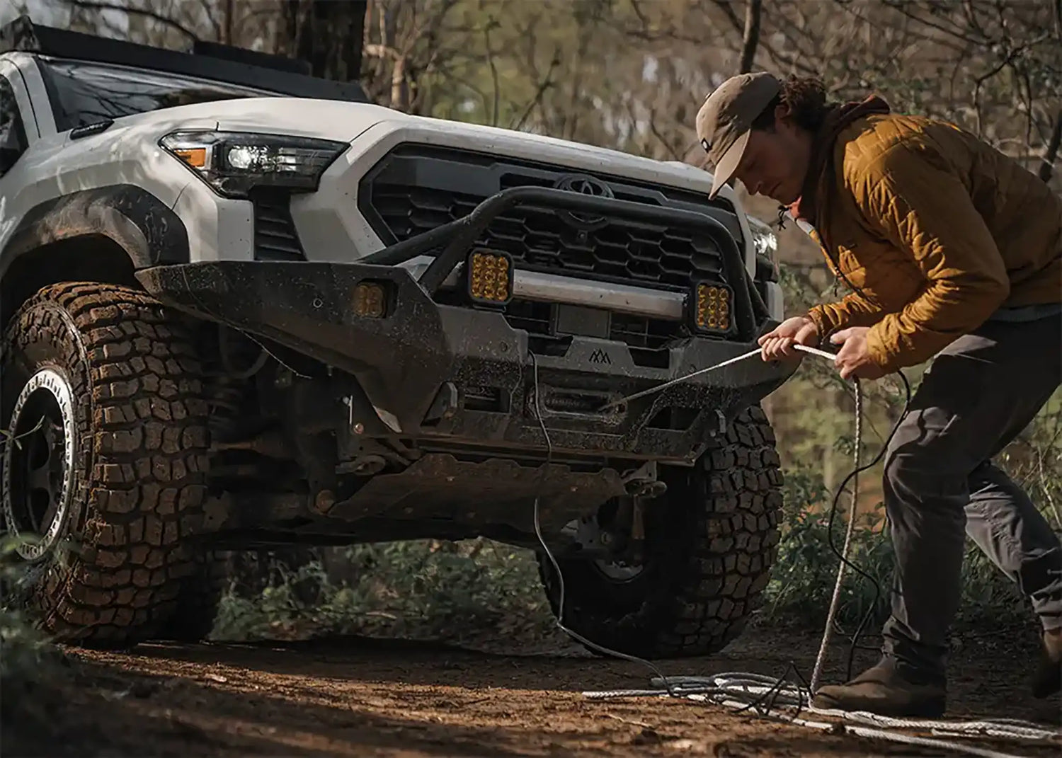 Person pulling line from a winch mounted to a Backwoods Adventure Mods 4x4 off-road bumper on an overland vehicle in a forested setting