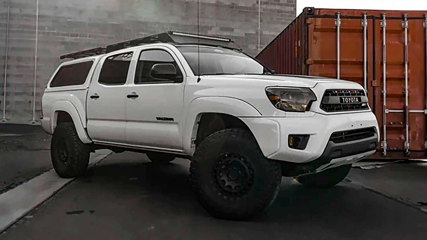 A white 2nd Gen Toyota Tacoma equipped with 2nd Gen Tacoma mods and accessories from Backwoods Adventure Mods, parked in front of a gray brick wall and an orange shipping container.