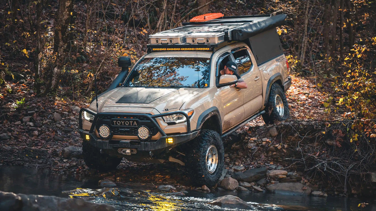 A beige 3rd Gen Toyota Tacoma equipped with rock sliders, bumpers, roof rack, bed rack, and other 3rd gen Tacoma mods from Backwoods Adventure Mods, driving through a rocky stream off road in a wooded setting.