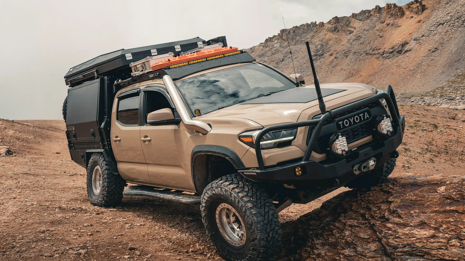 Tan 3rd Gen Toyota Tacoma driving off road through rocky desert terrain with a 3rd Gen Tacoma Roof Rack and various roof rack accessories from Backwoods Adventure Mods