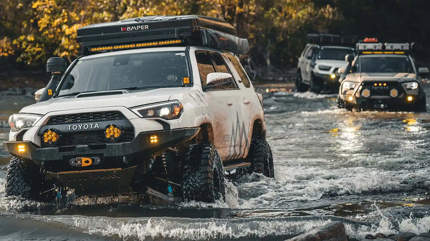 A white 5th Gen 4Runner equipped with mods and accessories from Backwoods Advenrture Mods driving through a deep water crossing off road leading other overland toyota vehicles visible in the background