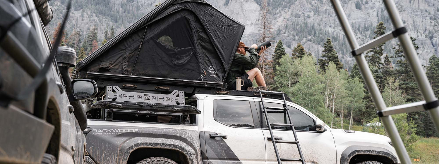Image of a man sitting on a Toyota Tacoma roof rack with with a roof top tent mounted behind him on top of the bed rack.