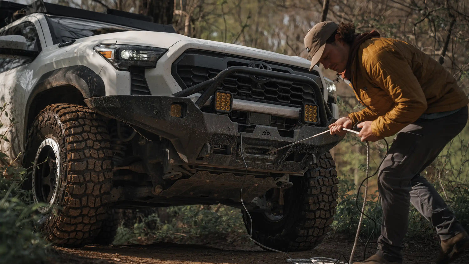 A man uses an off-road winch from Backwoods Adventure Mods installed on the front bumper of a white Toyota Tacoma overland truck with large mud-covered tires, to perform a vehicle recovery on a forest trail.