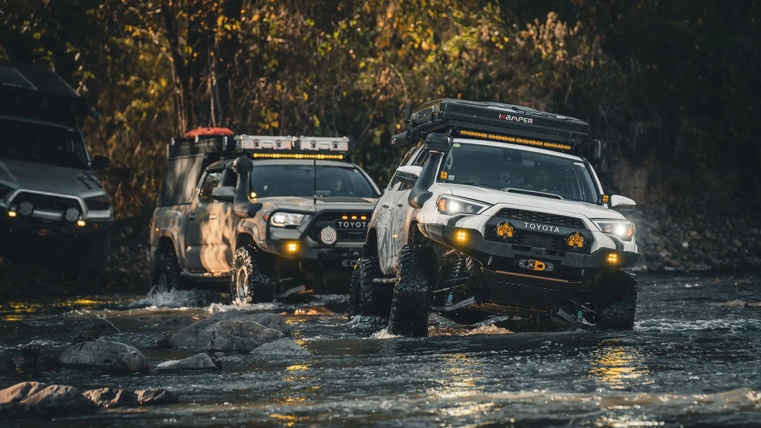 Three off-road vehicles, equipped with Backwoods Adventure Mods Offroad Lighting solutions, navigating a rocky river crossing in a forest at dusk