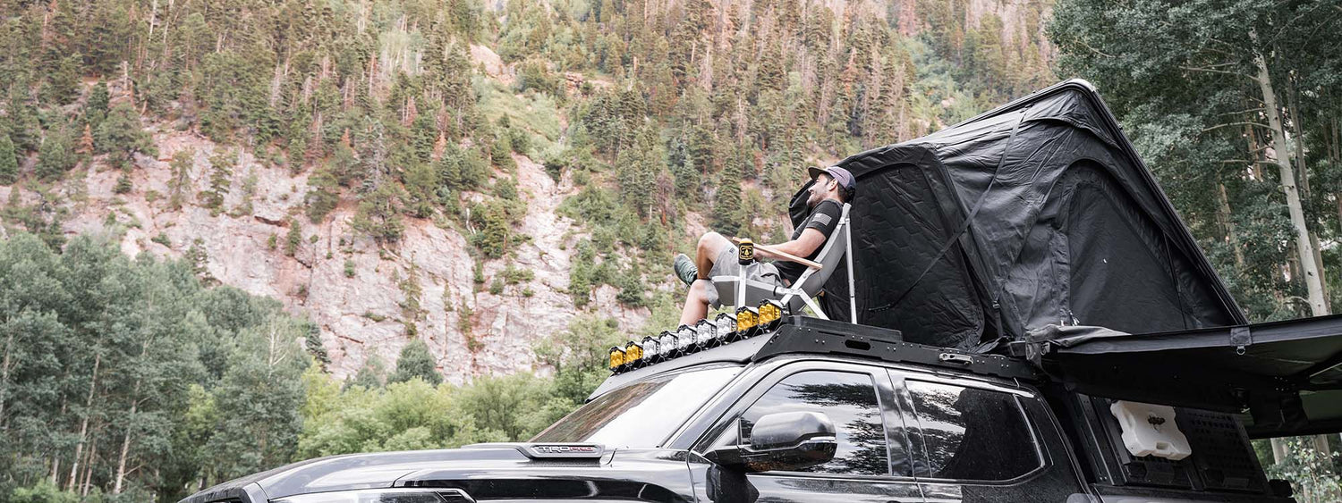 A new toyota tundra camping in the colorado mountains with a backwoods adventure mods driftr roof rack and bed rack. A man is sitting in a camp chair on top of the roof rack looking at the views.