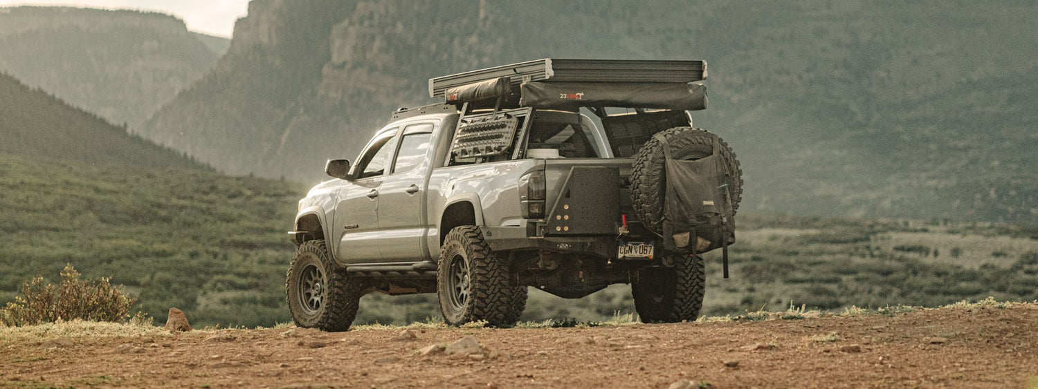 A 2018 toyota tacoma with a backwoods adventure mods rear bumper parked at sunset camping outdoors.