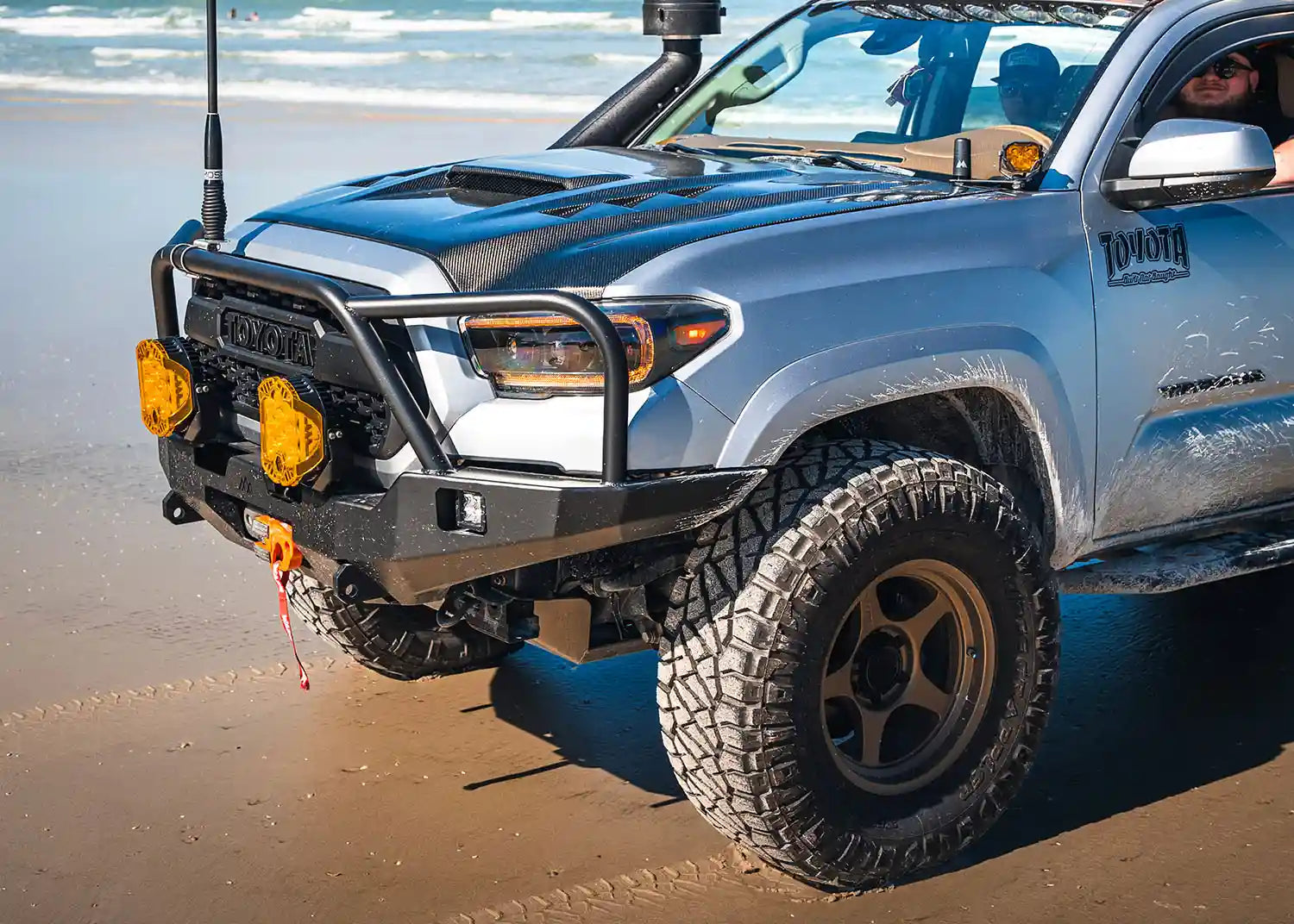 2016-2023 Toyota Tacoma Hi-Lite Overland Front Bumper with Bull Bar from Backwoods Adventure Mods installed on a light blue 3rd Gen Tacoma driving on the beach