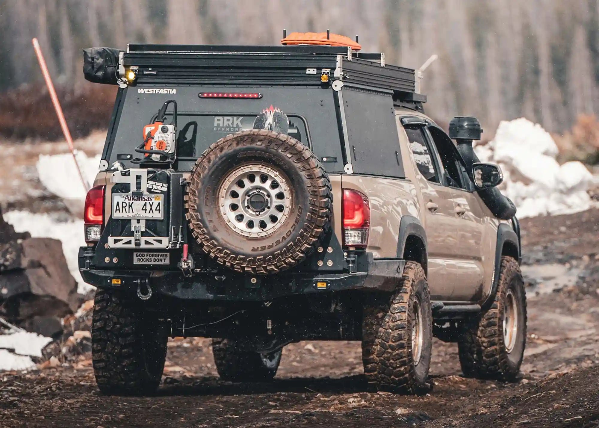 Rear view of a 3rd Gen Toyota Tacoma with a Backwoods Adventure Mods High Clearance Dual Swing Out Rear Bumper installed and a spare tire mounted on the bumper, driving off road in through the mud
