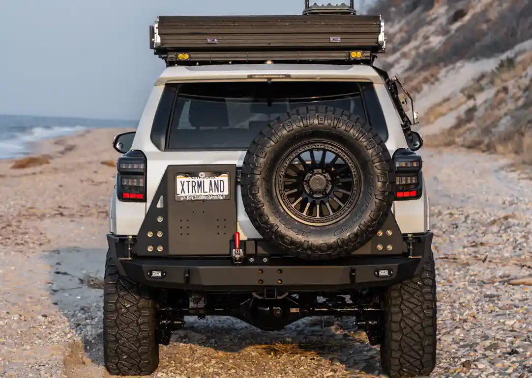 Rear view of a 5th Gen Toyota 4Runner with a Backwoods Adventure Mods high clearance dual swing out bumper and spare tire carrier drivning off road on a beach