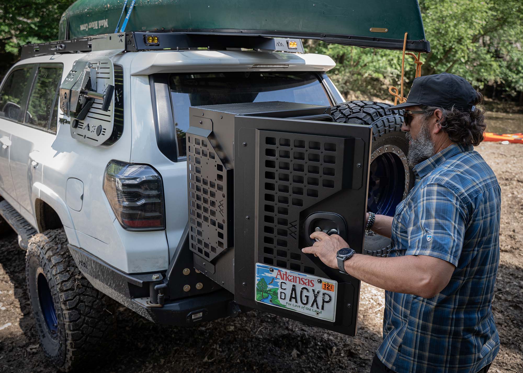 Man opening the Backwoods Adventure Mods Overland Storage Box with exterior molle panels visible. The storage box is mounted on the rear of a white overland built Toyota 4Runner with a canoe mounted to the roof rack.