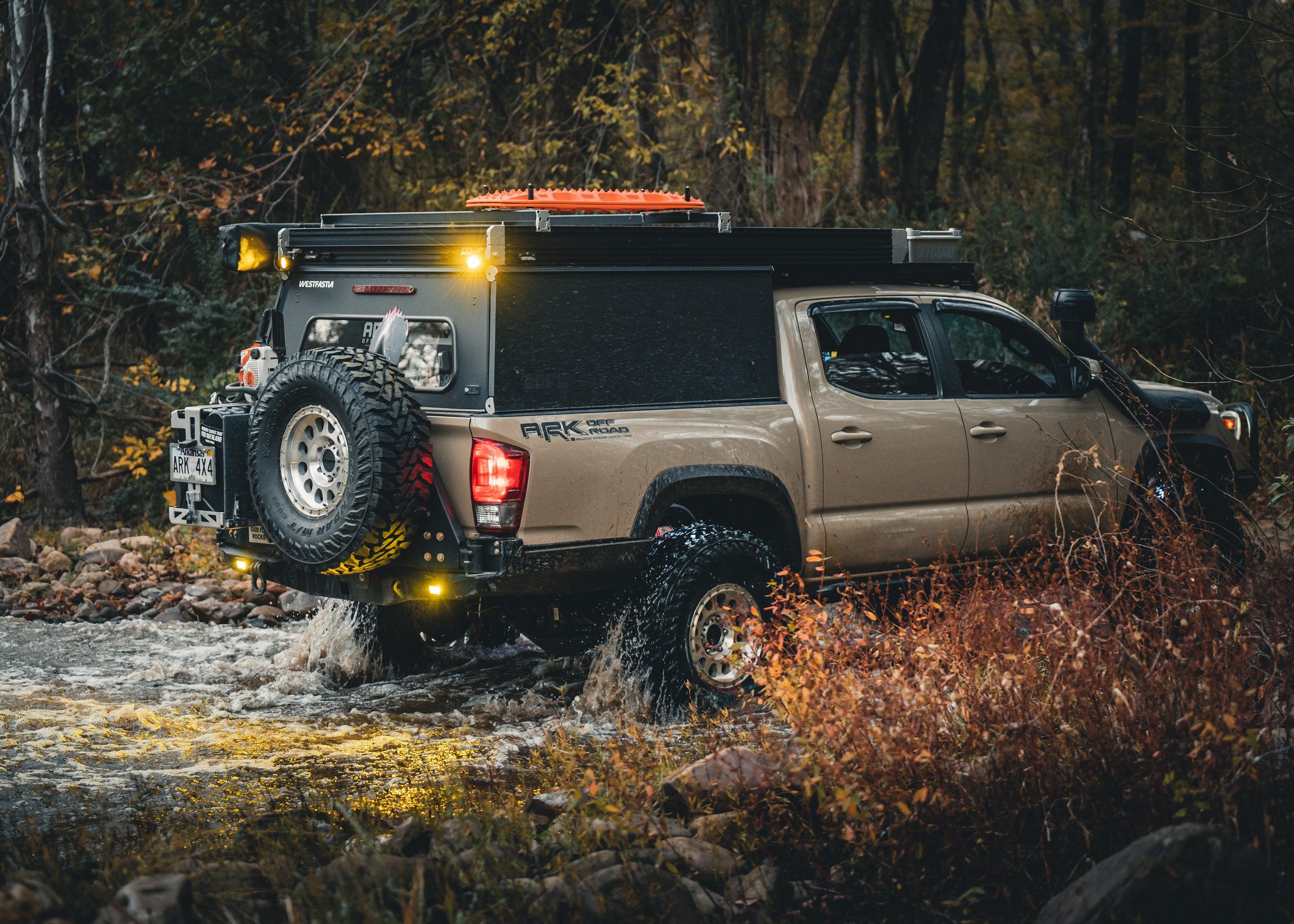 Backwoods Adventure Mods 2016-2023 Toyota Tacoma Hi-Lite High Clearance Dual Swing Out Rear Bumper installed on a tan 3rd Gen Tacoma driving off road through a water crossing