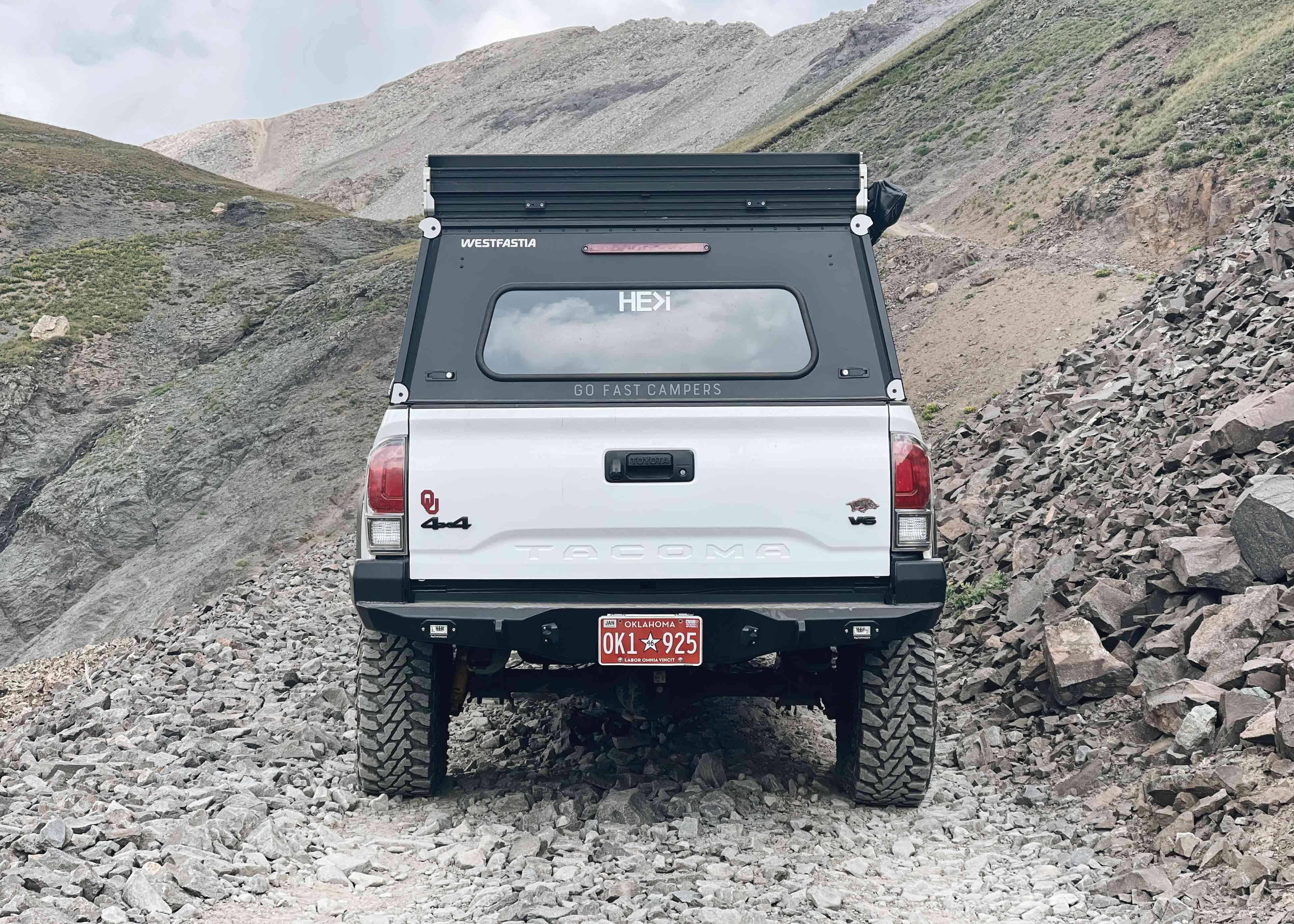 Rear-facing view of a White 3rd Gen Toyota Tacoma driving off road in the mountains with a Backwoods Adventure Mods Hi-Lite High Clearance Rear Bumper installed 